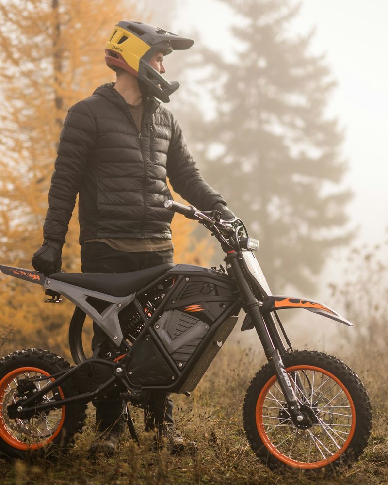 A rider in a full-face helmet standing beside an electric off-road motorcycle in autumn fog