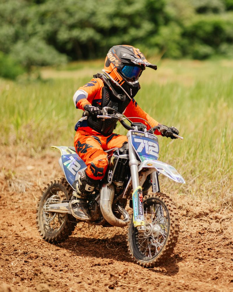 A young rider in full kit on a small off-road motorcycle on a grass and earth track